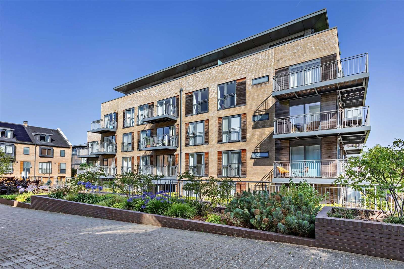 Modern residential apartment building with brick façade, multiple balconies with metal railings, and landscaped grounds featuring shrubs and plants.